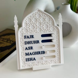 May include: A white prayer time tracker with an Islamic design. The tracker features five slots labelled FAJR, DHUHR, ASR, MAGHRIB, and ISHA, each with a blue slider. The design includes a decorative cutout pattern.