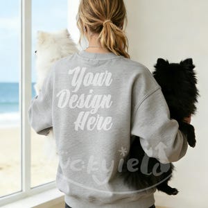 Könnte beinhalten: Ein hellgraues Sweatshirt mit dem Text "Your Design Here" in weißer Schrift auf dem Rücken. Eine Person hält einen weißen und einen schwarzen Hund. Die Hunde sind flauschig und der Hintergrund ist ein Fenster mit Blick auf das Meer.