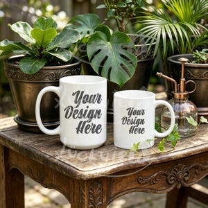 May include: Two white ceramic mugs with the text "Your Design Here" are displayed on a wooden table. The mugs are different sizes, with a large mug and a smaller mug. The background includes potted plants and a glass spray bottle.