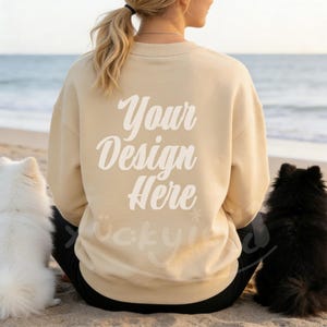 May include: A beige sweatshirt with the text "Your Design Here" in white script on the back. The sweatshirt is worn by a person sitting on a sandy beach with two dogs, one white and one black, on either side.