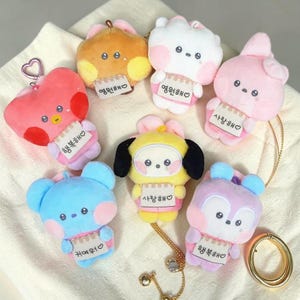 May include: A collection of plush keychains in various colors, including pink, blue, yellow, and orange. Each keychain features a cartoon character design with a heart-shaped face and a small tag with Korean text. A gold-colored chain and ring are also visible.