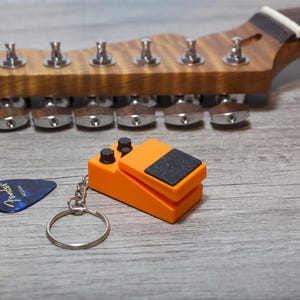 以下が含まれることがあります： 黒いディテールとシルバーのキーリングが付いたオレンジ色のギターペダルキーホルダー。白い文字が入った青いギターピックがキーホルダーの隣にあります。背景には、シルバーのチューニングペグが付いた木製のギターヘッドストックがあります。