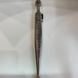 May include: A silver dagger with an ornate sheath. The dagger and sheath are covered in detailed floral and geometric patterns. The handle has a rounded top with a small design. The sheath has a pointed end.