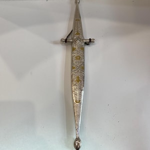 May include: A silver dagger with ornate gold and floral designs. The dagger has a pointed blade and a decorative handle with a small guard. A brown leather strap is attached to the handle.