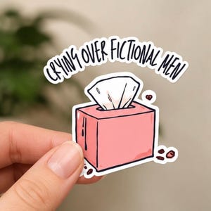 May include: A pink sticker featuring a cartoon tissue box with tissues and the text "CRYING OVER FICTIONAL MEN" in a black handwritten font. The sticker is held by a person's hand.