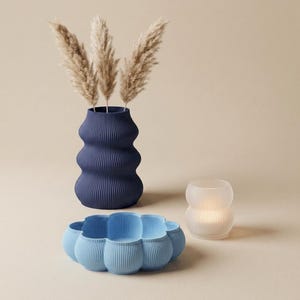 Puede incluir: Un jarrón acanalado de color azul oscuro que contiene tres tallos de hierba de la pampa beige, un cuenco acanalado de color azul claro con borde festoneado y un pequeño portavelas blanco translúcido acanalado con luz cálida.