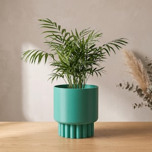 May include: A vibrant teal cylindrical plant pot with a smooth upper section and a vertically fluted, ribbed base. The pot contains a lush green parlour palm plant with multiple feathery fronds extending upwards from the centre.