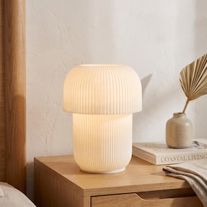 May include: A white table lamp with a mushroom-shaped silhouette and a vertically ribbed texture. The lamp emits a soft, warm glow from within its cylindrical base and rounded shade, creating a uniform, light-diffusing effect across the surface.