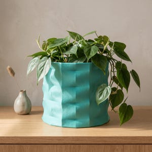 May include: A teal cylindrical planter with a vertical ribbed texture containing a trailing green heart-leaf philodendron plant. Beside the planter sits a small, light-coloured ceramic bud vase holding a single dried botanical stem.