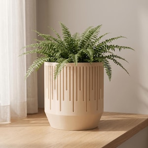 May include: A light beige cylindrical planter featuring a vertical ribbed texture with varying lengths of recessed lines around the exterior. A lush green fern with delicate, feathery fronds is planted inside, extending outward from the top rim.
