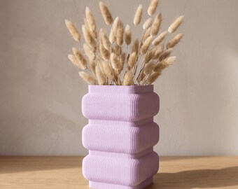 May include: A light purple vase with a ribbed, three-tiered square structure. The vase is filled with a bouquet of dried, fluffy beige bunny tail grass stems that extend upwards from the top opening of the container.