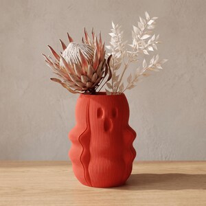 May include: A vibrant orange vase with a distinct vertical ribbed texture and a rounded, undulating silhouette. The vase contains a large, dried pale pink protea flower and several sprigs of dried white foliage, creating a floral arrangement.