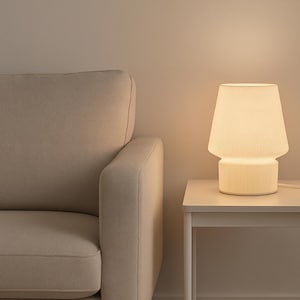 Puede incluir: Una lámpara de mesa blanca con pantalla y base acanaladas, que emite un brillo cálido. La lámpara está sobre una pequeña mesa auxiliar blanca junto a un sofá beige. La escena está iluminada por la suave luz de la lámpara, creando un ambiente acogedor.