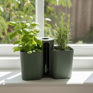May include: A dark green, dual-compartment planter with a ribbed texture and a central vertical glass tube. The left side contains a leafy green basil plant, while the right side holds a sprig of rosemary, both positioned on a white windowsill.