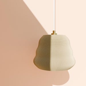 May include: A beige, wavy-shaped pendant light with a textured surface. It hangs from a white cord and a gold-coloured fixture. The background is a solid peach colour, with a shadow cast on the wall.