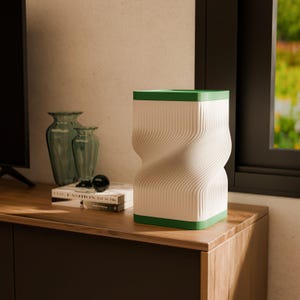 May include: A white and green decorative vase with a unique, twisted design. The vase has a rectangular shape with a green top and bottom. The body of the vase features vertical, wave-like lines. The vase is sitting on a wooden surface.