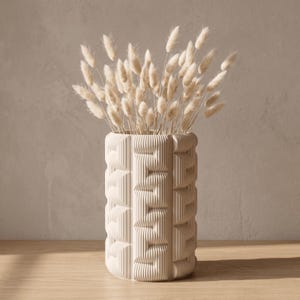 May include: A cylindrical off-white vase with a repeating vertical ribbed texture and geometric indentations. The vase is filled with a bouquet of dried, fluffy, cream-colored bunny tail grass stems, creating a soft and neutral display.