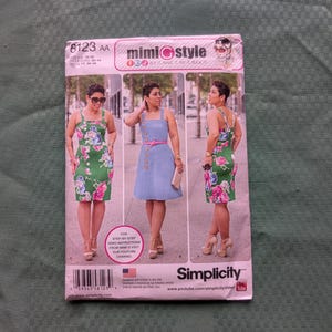 May include: A Simplicity sewing pattern envelope 8123 for a Mimi G Style dress. The front displays three views of a sleeveless dress with a square neckline, featuring a green floral print version and a solid light blue version with buttons.
