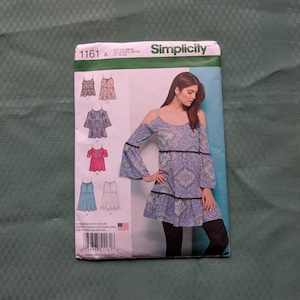 May include: A Simplicity sewing pattern envelope 1161 featuring illustrations of four different top designs. The main image shows a blue and purple paisley print cold-shoulder tunic with bell sleeves and black trim, sized for US XS-XL.