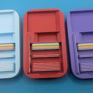 Op de afbeelding: Drie plastic opbergcontainers met deksels, elk in een andere kleur: lichtblauw, rood en paars. Elke container heeft drie niveaus met regenboogkleurige scheidingswanden.