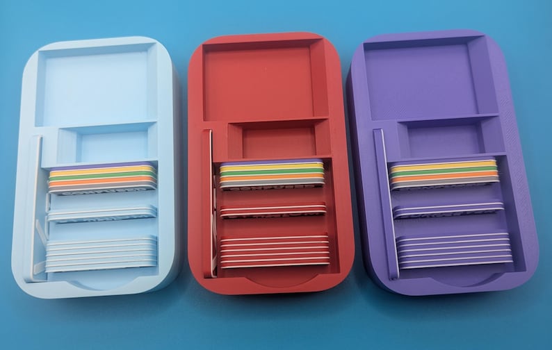 Op de afbeelding: Drie plastic opbergcontainers met deksels, elk in een andere kleur: lichtblauw, rood en paars. Elke container heeft drie niveaus met regenboogkleurige scheidingswanden.