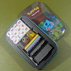 May include: A metal tin with a Pokemon Trading Card Game design. The tin is open and contains a set of dice, a deck of cards, and a few other accessories.