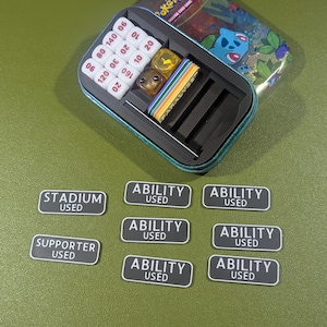 Op de afbeelding: Een groene en blauwe metalen doos met een Pokemon Trading Card Game logo en een cartoon afbeelding van een blauwe Pokemon. De doos bevat een set dobbelstenen, waaronder een gele d6, een bruine d6 en een witte d10. De doos bevat ook een set zwart-witte tokens die "Stadium Used", "Supporter Used" en "Ability Used" zeggen.