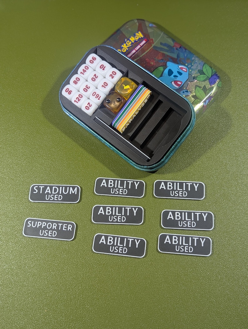 Op de afbeelding: Een groene en blauwe metalen doos met een Pokemon Trading Card Game logo en een cartoon afbeelding van een blauwe Pokemon. De doos bevat een set dobbelstenen, waaronder een gele d6, een bruine d6 en een witte d10. De doos bevat ook een set zwart-witte tokens die "Stadium Used", "Supporter Used" en "Ability Used" zeggen.