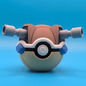 May include: A brown and beige Poke Ball with two gray cannons on the sides. The Poke Ball has a white rim and a blue and white design on the front.