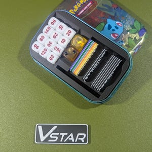 Op de afbeelding: Een blauwe en groene tin met het Pok&eacute;mon Trading Card Game-logo. De tin is open en bevat een set dobbelstenen, een set tellers en een set kaarten. De tin staat op een groene ondergrond.