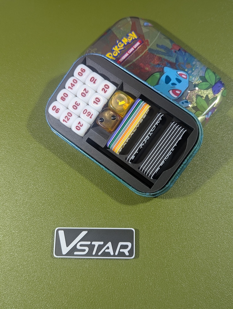Op de afbeelding: Een blauwe en groene tin met het Pok&eacute;mon Trading Card Game-logo. De tin is open en bevat een set dobbelstenen, een set tellers en een set kaarten. De tin staat op een groene ondergrond.