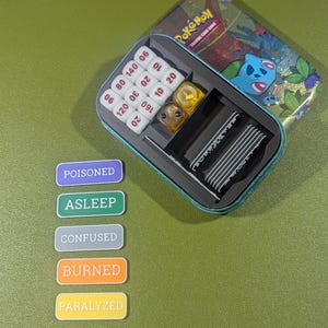 Op de afbeelding: Een blauwe en groene tinnen doos met het woord "Pok&eacute;mon" erop. De doos is open en bevat een set dobbelstenen, een set conditie markers en een set kaarten. De conditie markers zijn gelabeld "Vergiftigd", "Slaap", "Verward", "Verbrand" en "Verlamd".