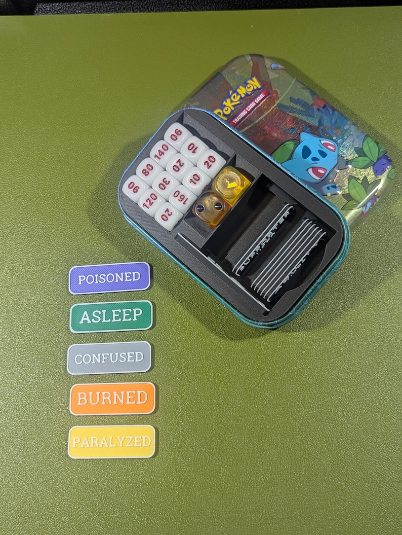 Op de afbeelding: Een blauwe en groene tinnen doos met het woord "Pok&eacute;mon" erop. De doos is open en bevat een set dobbelstenen, een set conditie markers en een set kaarten. De conditie markers zijn gelabeld "Vergiftigd", "Slaap", "Verward", "Verbrand" en "Verlamd".