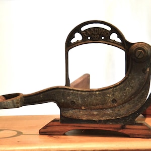 May include: A vintage Raadvad bread slicer featuring a dark, weathered cast iron frame with a curved blade and a wooden handle. The metal structure displays the brand name Raadvad with a crown emblem at the top, mounted on a wooden base.