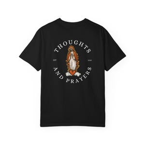 以下が含まれることがあります： 黒いTシャツ。円形のデザインで、「THOUGHTS AND PRAYERS」の文字が、心臓を持つ宗教的な人物のイラストを囲んでいます。グラフィックには「EST 2025」の文字も含まれています。
