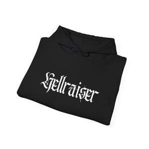 May include: Black hoodie with the word "Hellraiser" printed in white gothic lettering across the front. The hoodie has a drawstring hood and is folded neatly. The fabric appears to be a soft material.