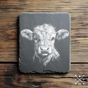 以下が含まれることがあります： 黒いスレートコースターに、白い線で描かれた子牛の顔のイラスト。子牛は正面を向いて見ている。