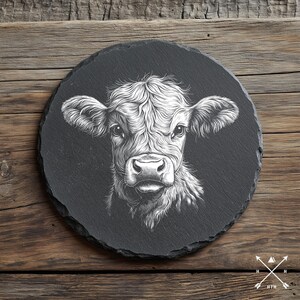 以下が含まれることがあります： 黒い円形の石板コースターに、子牛の顔の白黒のイラストが描かれています。