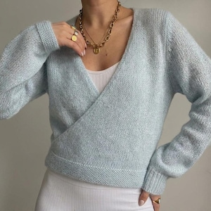 Könnte beinhalten: Ein hellblauer, langärmeliger Wickelpullover mit V-Ausschnitt und gerippten Bündchen, getragen über einem weißen Ripp-Unterhemd. Der Pullover hat eine weiche, flauschige Strickstruktur und eine kurze Passform, die auf der Taille endet.