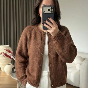May include: A brown, button-down cardigan with a round neckline. The cardigan is made of a textured knit material and has long sleeves. The buttons are a matching brown color. The person is wearing a white top and white pants.