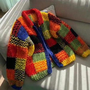 May include: A colorful, patchwork-style cardigan in a variety of vibrant colors including red, orange, yellow, green, blue, and black. The knitted sweater features a collared neckline and a blue closure. The garment is displayed on a light gray surface.