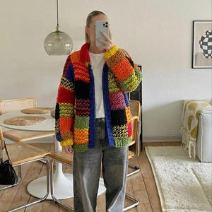 Può includere: Un cardigan colorato in stile patchwork nei toni del rosso, arancione, giallo, verde, blu e nero. Il maglione aperto ha un colletto e maniche lunghe. La persona indossa una maglietta bianca e jeans grigi.