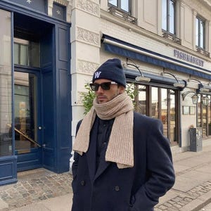 May include: A person wearing a navy blue beanie with a white "W" logo, sunglasses, a beige scarf, and a navy blue overcoat. The background includes a building with a "FISHMARKET" sign.