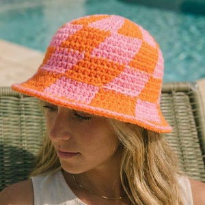 May include: A crochet bucket hat featuring a vibrant checkerboard pattern in alternating bright orange and soft pink yarn. The hat has a rounded crown and a downward-sloping brim, constructed with a visible textured stitch throughout the design.