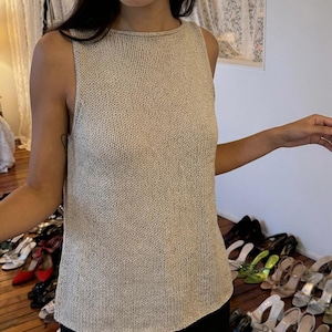 Peut inclure: Un haut sans manches en tricot beige avec un col rond. Le haut a un aspect texturé et un ourlet légèrement incurvé. L'arrière-plan présente une collection de chaussures sur un sol en bois.