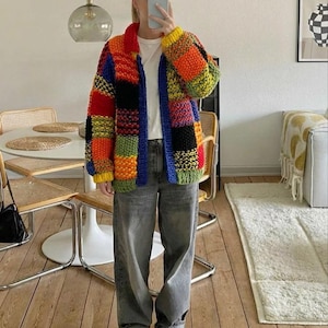 Könnte beinhalten: Eine grob gestrickte Strickjacke mit einem lebendigen Patchwork-Design aus roten, orangen, gelben, grünen, blauen und schwarzen Garnblöcken. Das Kleidungsstück hat einen Kragen, lange Ärmel mit gerippten Bündchen und eine Knopfleiste vorne.
