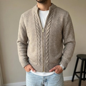 Può includere: Un cardigan beige con zip in maglia con un motivo a trecce sulla parte anteriore. Il cardigan ha un collo alto e polsini a coste. La cerniera è color oro. La persona indossa una maglietta bianca e jeans blu.