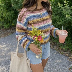 Può includere: Un maglione all'uncinetto a maniche lunghe con righe orizzontali nei toni del rosa, blu, giallo e marrone. Il maglione presenta un'ampia scollatura e polsini a coste. È abbinato a pantaloncini di jeans azzurri e una borsa in tela beige.
