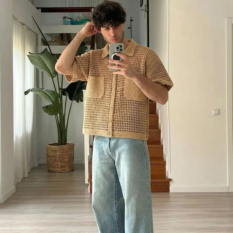 May include: A tan crochet shirt with short sleeves and two front pockets. The shirt is buttoned up and worn with light blue jeans. The person is holding a phone and taking a selfie. A large green plant is in the background.