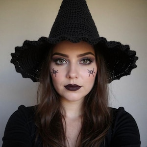 May include: A black crochet witch hat with a wide, ruffled brim. The hat is worn by a person with dark makeup, including dark lipstick and spider face paint. The person is wearing a black top.